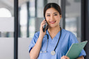 Nurse in blue scrubs using a phone and holding a binder clip, with a blurred healthcare office background, illustrating mobile device use in clinical settings